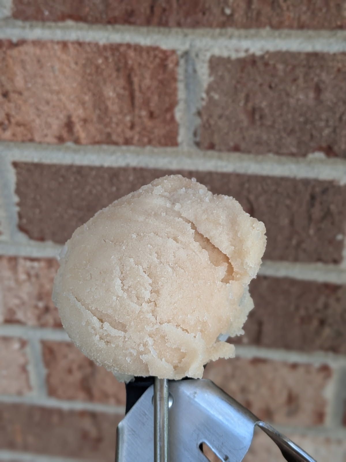 Close-up of a scoop of brown sugar held in metal tongs against a brick wall background.